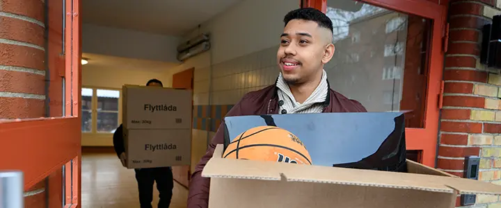Person bär en flyttlåda ut från ett flerbostadshus. En annan person bär flyttlådor i bakgrunden.